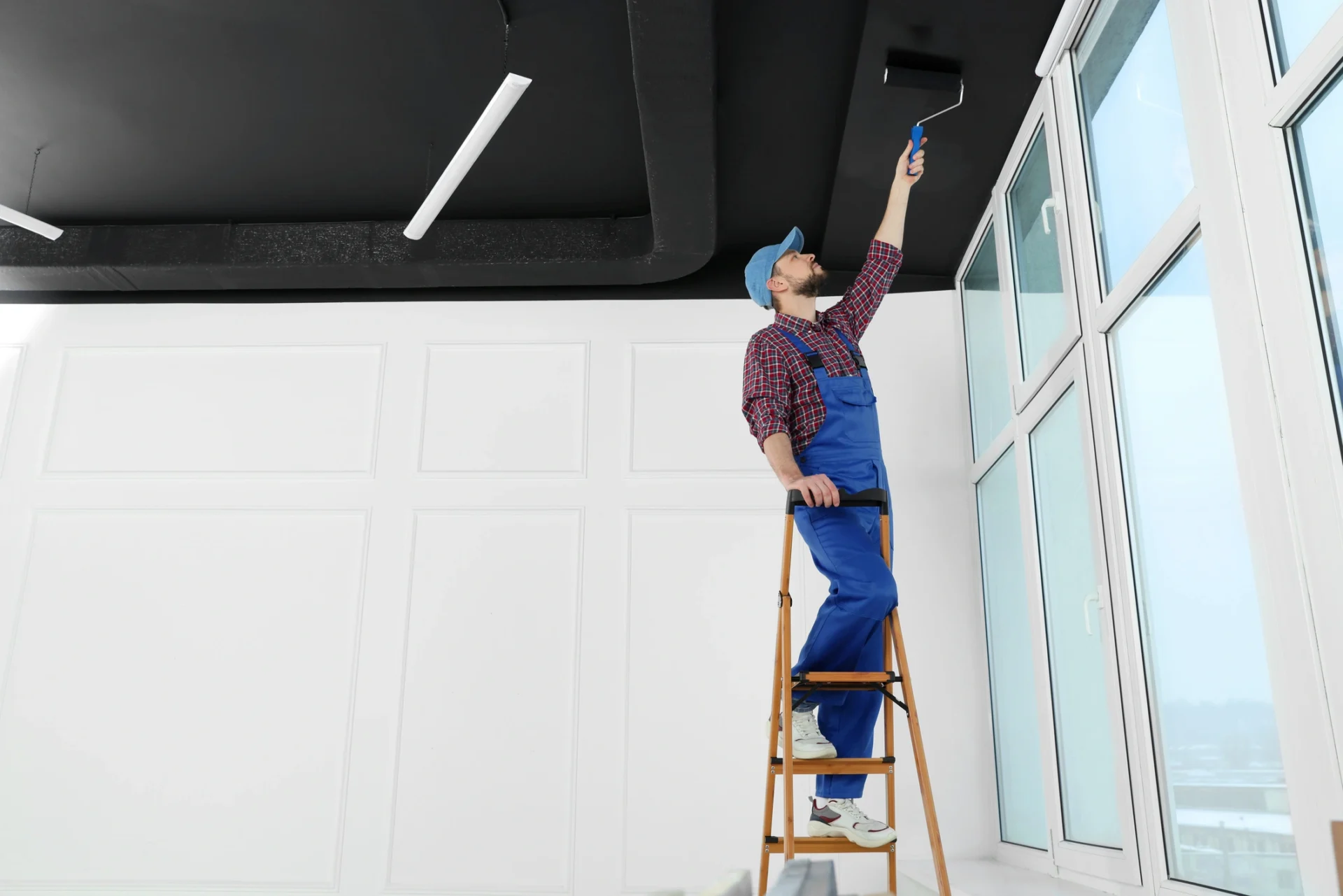 Worker in uniform painting ceiling with roller on stepladder indoors.